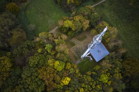 Das Luftbild zeigt einen großen Kommunikationsturm, der in einem bewaldeten Gebiet steht. Der Turm befindet sich neben einer kleinen Struktur mit einem grauen, schrägen Dach. Um den Turm und das Gebäude herum sind dichte Bäume in verschiedenen Grüntönen sowie Gelb- und Brauntönen zu sehen.