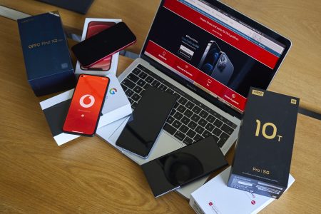 Das Bild zeigt einen Holztisch mit mehreren elektronischen Geräten und Boxen, die um einen geöffneten Laptop angeordnet sind. Der Laptop hat einen sichtbaren Bildschirm, der eine Webseite mit Smartphones anzeigt. Es sind mehrere Smartphone-Boxen vorhanden, darunter solche mit der Aufschrift „Oppo Find X2“ und „10T Pro 5G“. Verschiedene Smartphones, einige mit sichtbaren Bildschirmen und andere ausgeschaltet, sind auf den Boxen und dem Tisch platziert.