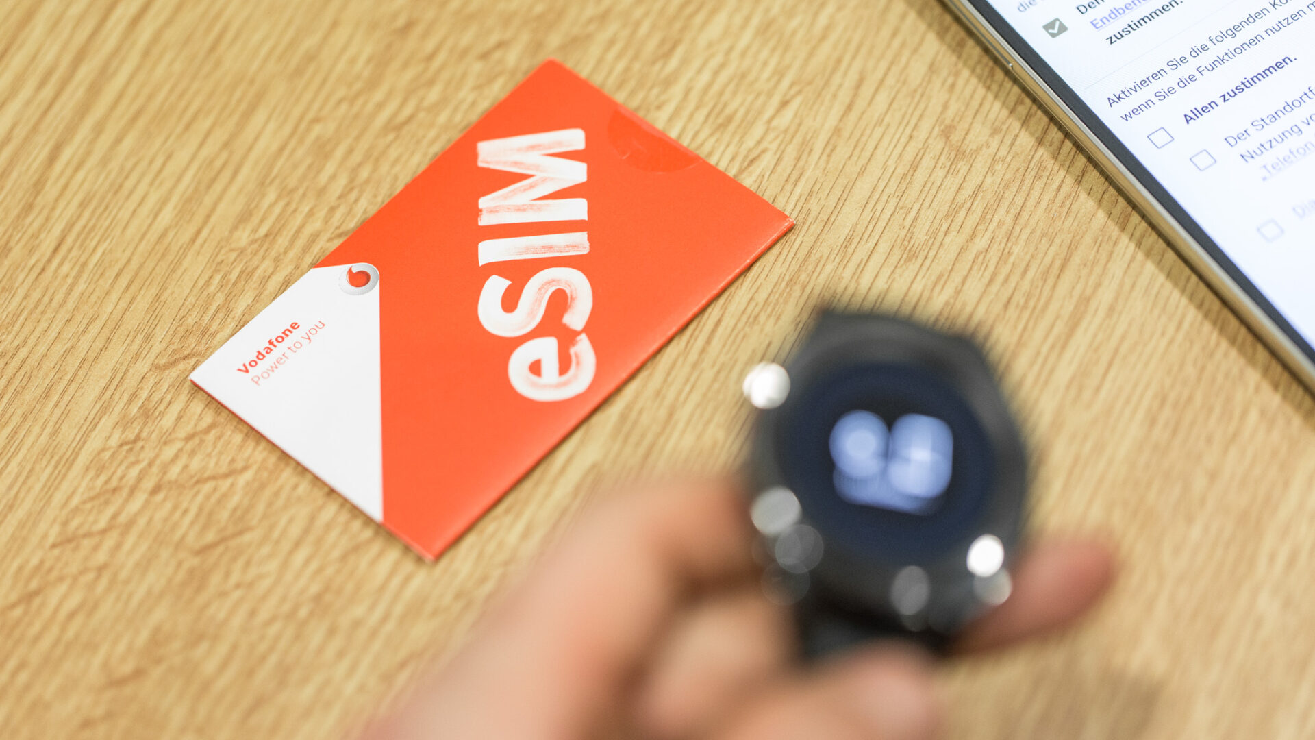 Auf dem Bild liegt eine rot-weiße Karte mit der Aufschrift „eSIM“ gut sichtbar auf einer hellen Holzoberfläche. Im Vordergrund hält eine unscharfe Hand eine schwarze Smartwatch, deren Display aktive Symbole zeigt. Rechts oben im Bildrand ist außerdem ein Smartphone zu erkennen, auf dessen Display eine App oder Webseite mit Checkboxen und Auswahlmöglichkeiten geöffnet ist.
