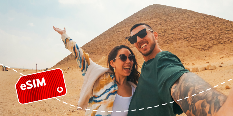 Zwei Personen posieren vor einer Pyramide und machen ein Selfie. Im Vordergrund ist ein rotes Etikett mit der Aufschrift „eSIM“ und dem Vodafone-Logo zu sehen.
