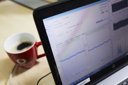 Ein Laptop-Bildschirm zeigt eine Software-Oberfläche mit Diagrammen und Datenanalysen von Nokia. Neben dem Laptop steht eine rote Tasse mit dem Vodafone-Logo, gefüllt mit Kaffee. Der Tisch hat eine helle Holzoberfläche, die eine professionelle Arbeitsumgebung vermittelt.