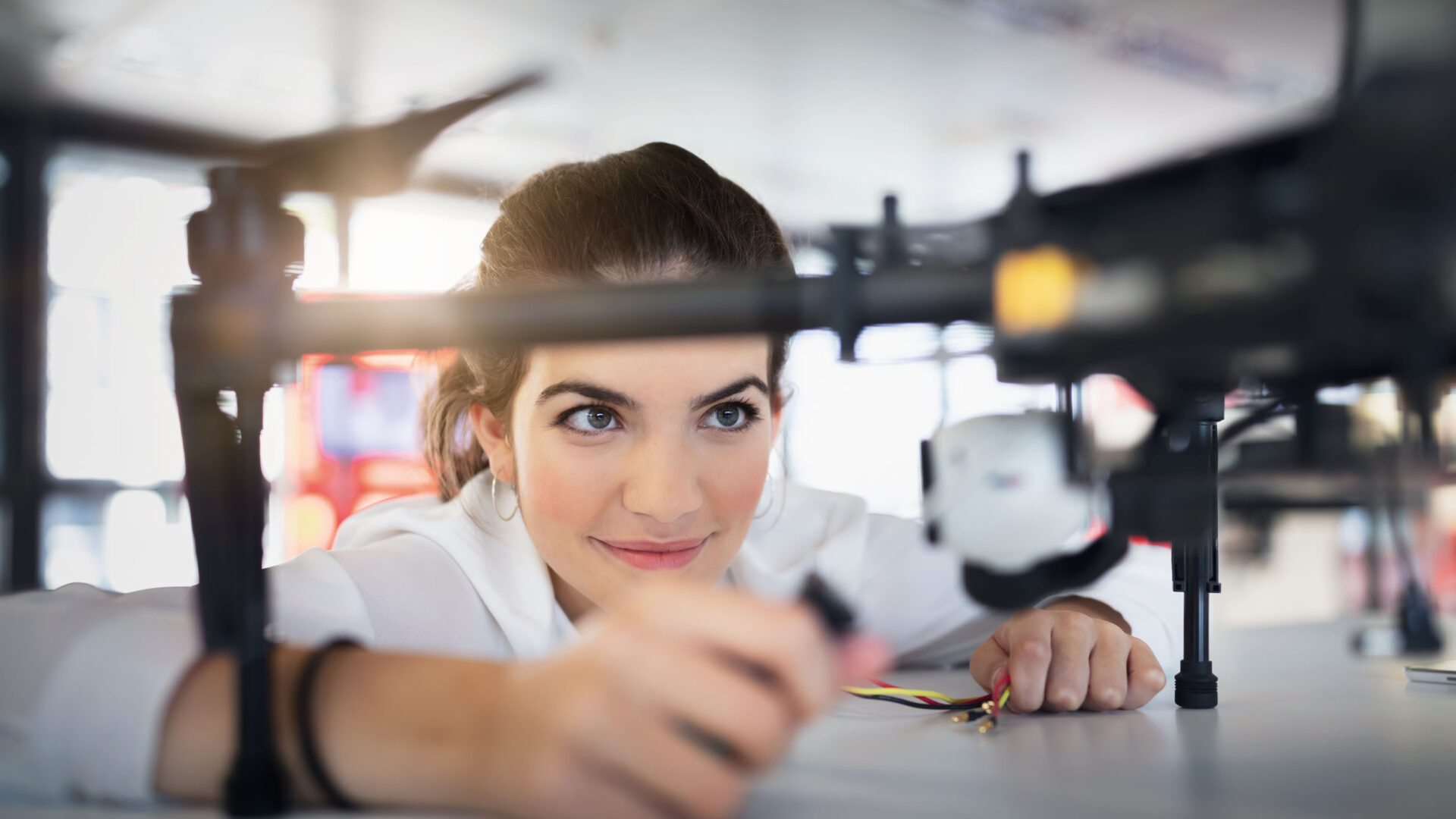 Eine Person arbeitet an einer Drohne auf einem Tisch. Die Drohne ist groß und hat mehrere Rotoren. Im Hintergrund sind verschwommene Details eines hellen Raumes zu sehen. Die Person hält ein kleines Werkzeug oder Bauteil in der Hand.