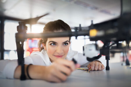 Eine Person arbeitet an einer Drohne auf einem Tisch. Die Drohne ist groß und hat mehrere Rotoren. Im Hintergrund sind verschwommene Details eines hellen Raumes zu sehen. Die Person hält ein kleines Werkzeug oder Bauteil in der Hand.