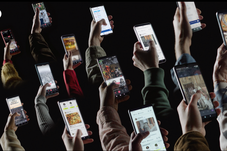 Mehrere Menschen halten ihre Smartphones in die Luft, wobei jeweils nur ihre Arme und Hände zu sehen sind. Die Bildschirme zeigen verschiedene Inhalte wie Social Media Posts, Chatnachrichten, Fotos und Webseiten. Die Personen tragen unterschiedliche Kleidung in Farben wie Rot, Beige und Grün. Der Hintergrund ist dunkel, sodass der Fokus auf den beleuchteten Bildschirmen und Händen liegt.