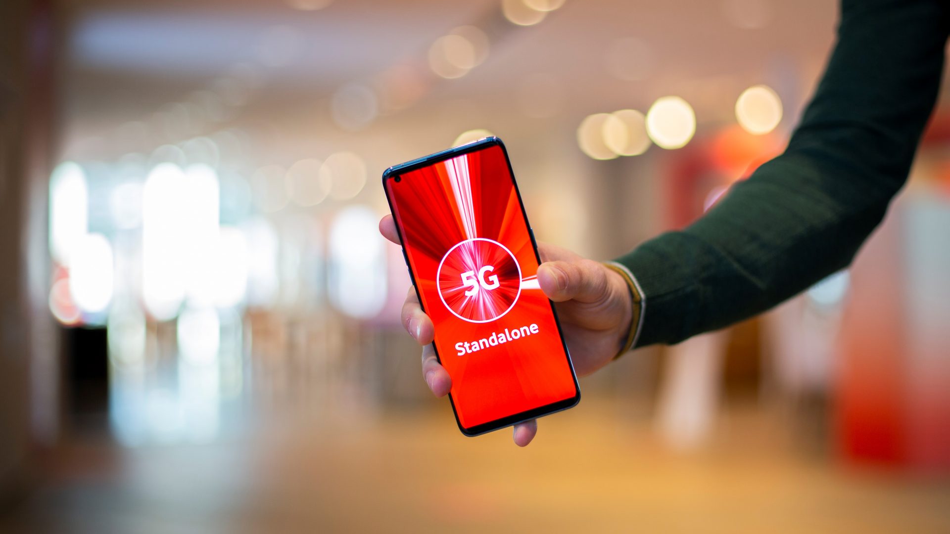Eine Hand hält ein Smartphone mit einem roten Bildschirm, auf dem "5G Standalone" steht. Der Hintergrund ist unscharf und zeigt einen Innenraum mit warmem Licht und modernen Einrichtungselementen.