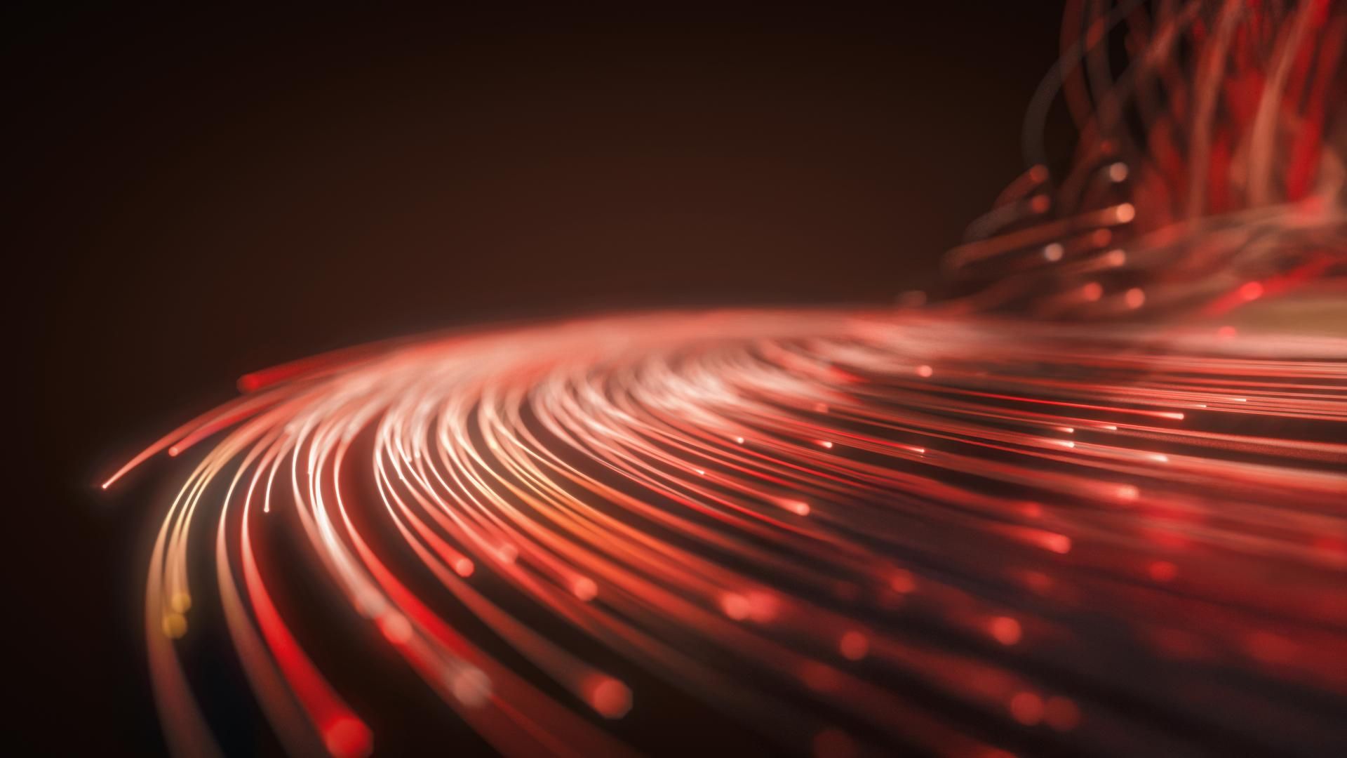 Das Bild zeigt eine Nahaufnahme von Glasfaserkabeln, die leuchtenden Rot strahlen. Die Kabel sind in Bündeln angeordnet und ihre Enden leuchten intensiv, was auf die Übertragung von Lichtsignalen hinweist. Der Hintergrund ist dunkel, wodurch die Farben der Glasfaserkabel besonders hervorgehoben werden. Diese Darstellung vermittelt den Eindruck von moderner Technologie und schneller Datenübertragung.