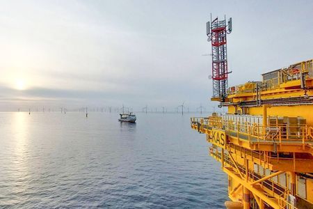 Eine gelbe Offshore-Plattform mit der Aufschrift "KAS" steht im Meer, neben einer roten und weißen Funkantenne. Im Hintergrund sind mehrere Windkraftanlagen und ein Schiff zu sehen, die eine weitläufige maritime Landschaft bilden. Der Himmel ist bewölkt und die Sonne scheint durch die Wolken.